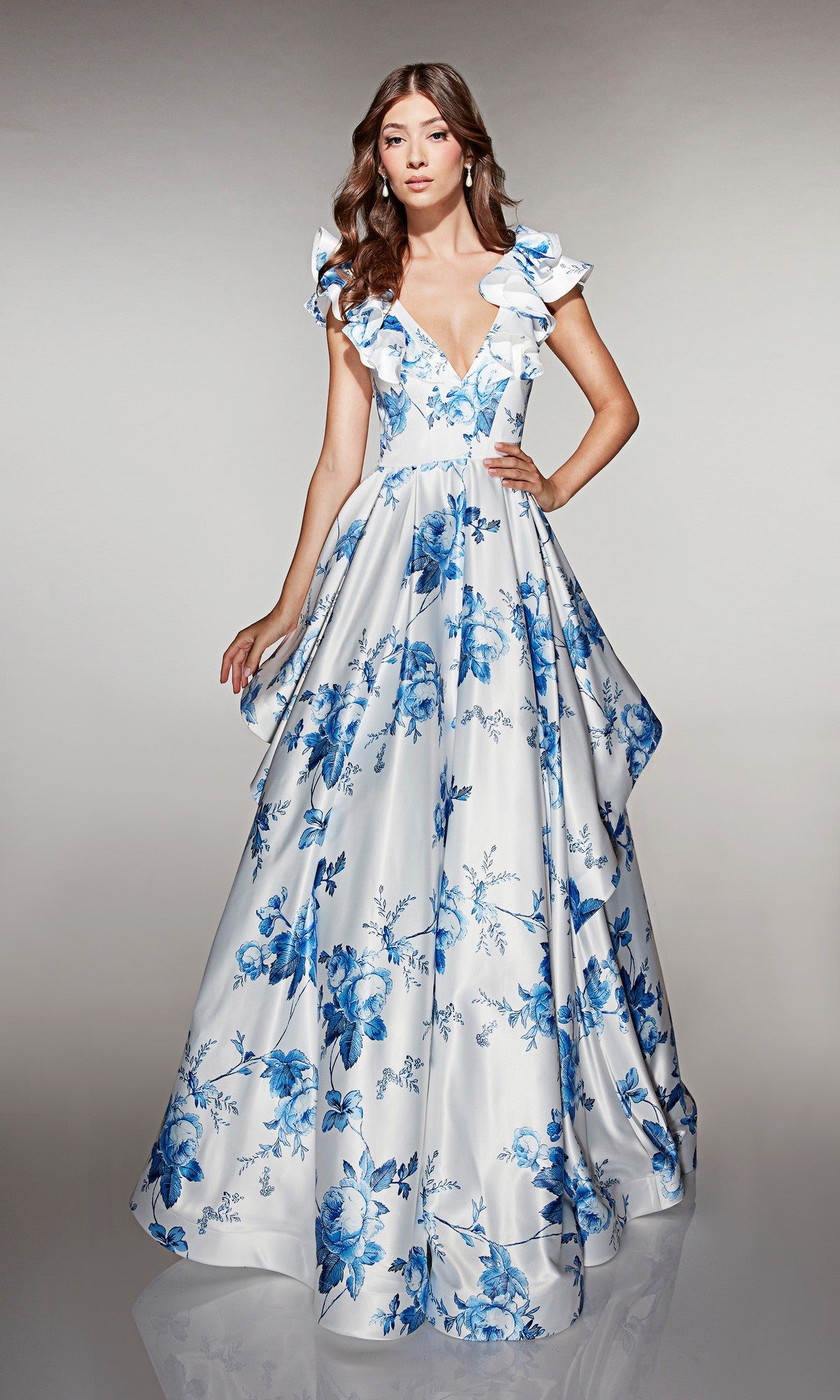 A woman poses in the Alyce 62001 long prom dress, a floor-length white satin gown with blue floral prints, ruffled cap sleeves, and a deep V neckline, set against a neutral background.